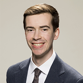 A photograph of staff member, Mark Jackson. He is wearing a navy suit and tie as he stands in front of a grey background and smiles at the camera.
